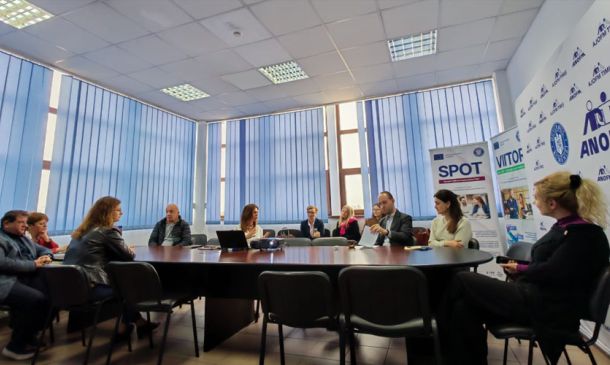People sit around a large conference table in a meeting room with blue blinds and banners along the wall