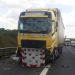 Yellow semi-truck with severe front-end damage on a highway, windshield cracked and debris scattered as traffic backs up nearby.