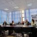 People sit around a large conference table in a meeting room with blue blinds and banners along the wall