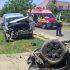Two damaged vehicles after a crash: a silver van with severe front-end damage on the grass and a flipped car on the road, with paramedics and ambulances nearby on a sunny day.