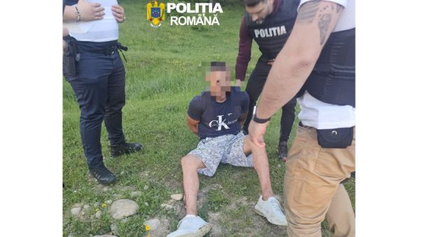 A man sits on grass with hands behind his back as three Romanian police officers surround him; face blurred, police logos visible above.
