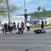 Crowd gathered at an intersection after a motorcycle crash; a rider lies on the pavement near a fallen bike while another motorcycle sits nearby and cars pass by.