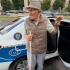 Older man with a cane and hat stands by the open rear door of a white and blue police car on a city street.