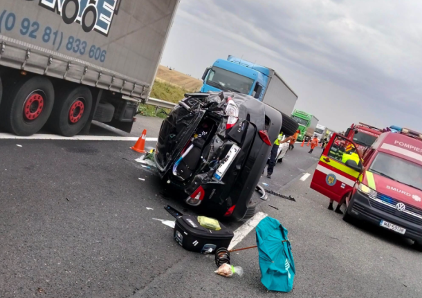 Upside-down car on a highway with emergency responders and a red ambulance nearby, cones marking the area.