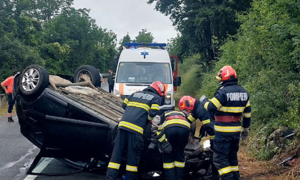Emergency responders work at a single-vehicle crash with an overturned car on a rural road, ambulance in background.