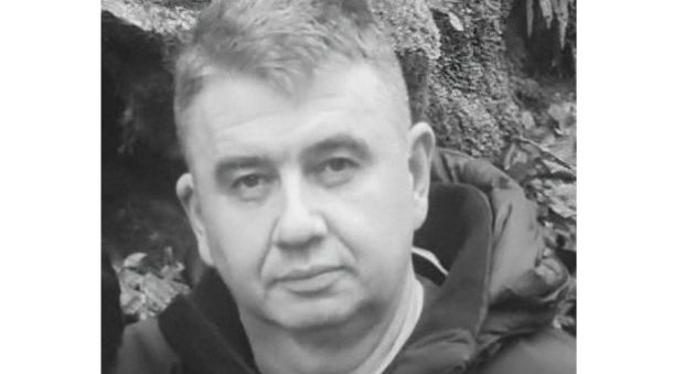 Black-and-white portrait of a middle-aged man with short hair wearing a dark jacket outdoors near rocks.