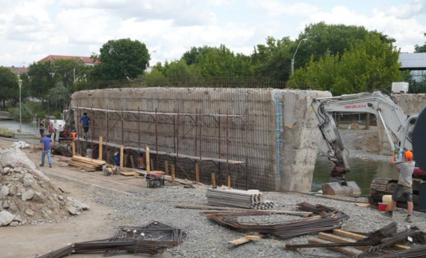 Construction site beside a river with a large reinforced concrete wall, rebar, and an excavator at work as workers move lumber and steel rods.