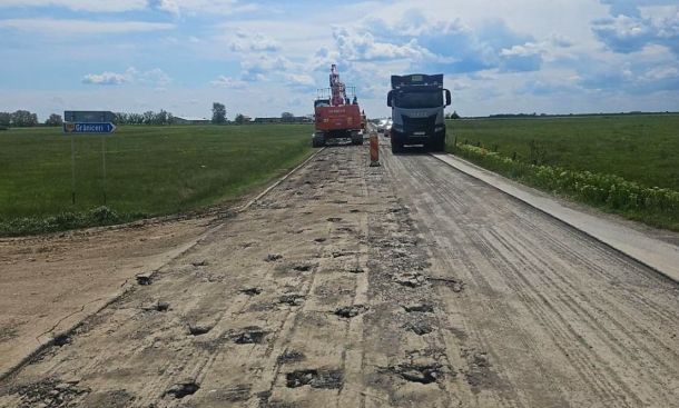 Rural road under repair with numerous potholes, heavy machinery (excavator) and a blue truck on the unpaved lane under a cloudy sky.