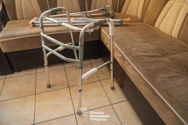 Folding aluminum walker with rubber tips set up in a tiled living room beside a beige sectional sofa.