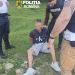 A man sits on grass with hands behind his back as three Romanian police officers surround him; face blurred, police logos visible above.