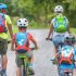 Four people (two adults and two children) ride bicycles on a rural path, wearing helmets and colorful backpacks.