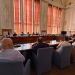 Formal meeting in a council chamber with people seated around a U-shaped wooden table and a large projector screen at the front.