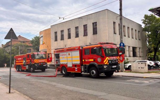 Two red fire trucks parked on a city street with hoses laid out near a beige multi‑story building under a cloudy sky, daytime scene.