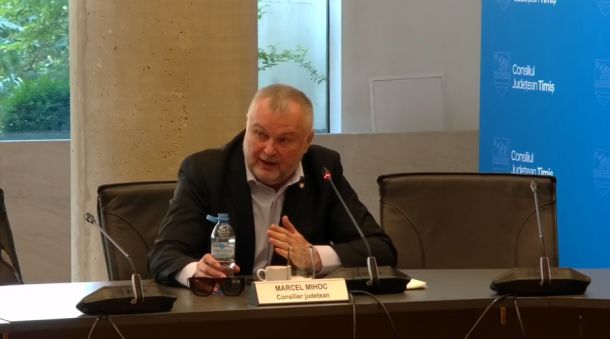 Man in a dark suit speaks into a microphone at a panel, nameplate reads 'Marcel Mihoc' and 'Consilier județean' beside a bottle of water on the table, with a blue council backdrop behind him