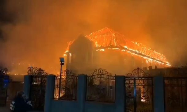House engulfed in flames at night, bright orange fire consuming the roof behind a decorative gate.