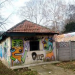 Small, graffiti-covered one-story building with a tiled, sloped roof and colorful cartoon faces on its walls, beside a path and fence.