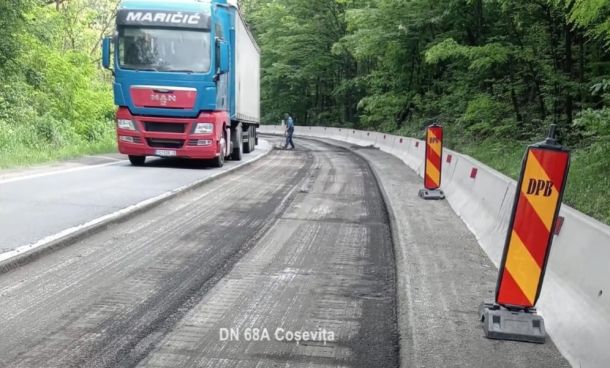 Blue and red semi-truck on a resurfacing road beside wooden barriers in a forested area.