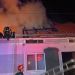 Firefighters on a burning building roof at night as flames and thick smoke rise, with a fire engine in view at the right. (Informative)