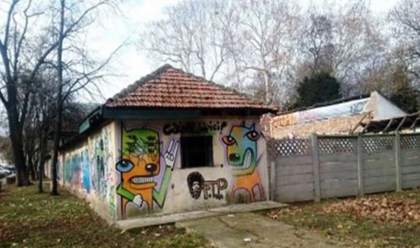 Small, graffiti-covered one-story building with a tiled, sloped roof and colorful cartoon faces on its walls, beside a path and fence.