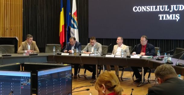 Five men sit at a long panel table in a council chamber, with flags behind and a large screen displaying 'CONSILIUL JUD TIMIȘ'.