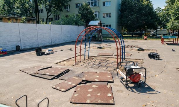 Playground under construction with red and blue looping arches, stacked mats, and construction equipment on a paved area.