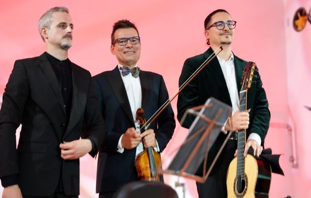 Three male musicians in suits on a pink-lit stage: one with a violin, one with a guitar, all looking toward the audience.