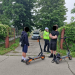 Two kids with orange-and-black scooters on a residential street as a police officer speaks nearby.