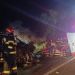 Night-time car crash scene with firefighters and a damaged black sedan on a highway, stretcher and medical crew ready for transport, emergency lights visible in the background