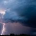 Lightning bolt streaks down through dark storm clouds over a flat horizon at dusk.