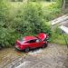 Red SUV sits in a shallow stream beneath a bridge as a rescuer attends to the driver, with onlookers leaning over the railing nearby.