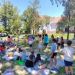 Large group of kids and adults in a sunny park, sitting on blankets and standing, participating in an outdoor activity.