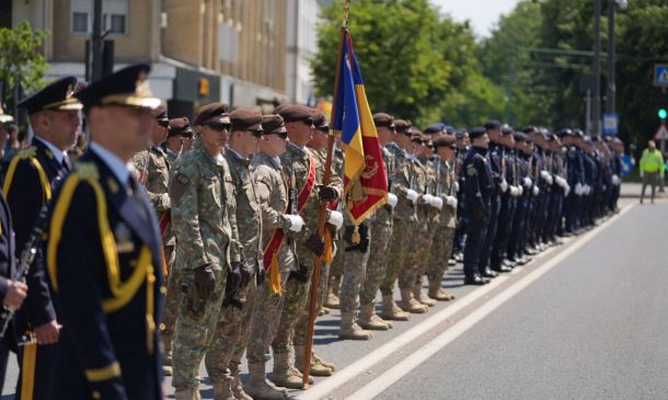 Line of soldiers in uniform standing at attention during a parade, one holding a blue-yellow-red flag.