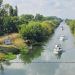 Boats cruising along a tree-lined canal with calm, reflective water viewed from above.