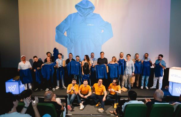 Group of people on a stage posing with blue jackets, in front of a large projected image of a blue hoodie on the screen behind them, an event photo showing staff and speakers holding up jackets.