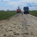 Rural road under repair with numerous potholes, heavy machinery (excavator) and a blue truck on the unpaved lane under a cloudy sky.