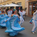 Group of dancers in flowing blue gowns performing a street parade, smiling and twirling feathers in the air against a city backdrop.