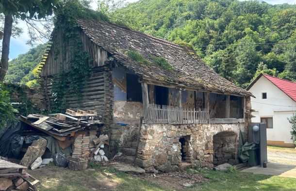 Old stone-and-wood cottage with a sagging roof, ivy growing on the walls, and a pile of lumber outside in a green hillside setting.