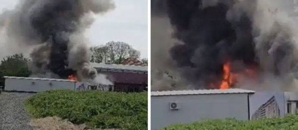 Industrial fire: thick black smoke and flames billow from a low-roof building near a gravel lot and trailers.