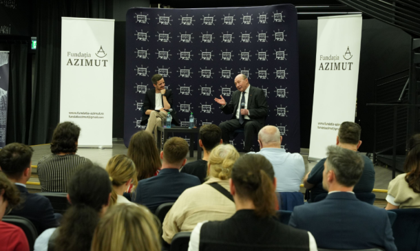 Two men on a stage in a panel discussion, audience seated in front, banners display Fundatia AZIMUT.