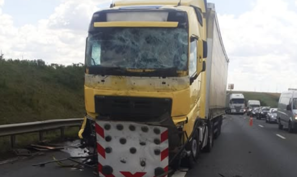 Yellow semi-truck with severe front-end damage on a highway, windshield cracked and debris scattered as traffic backs up nearby.