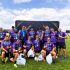 Rugby team in purple jerseys posing on a sunny field with medals and a trophy after winning.