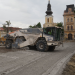 White road roller compactor on a dirt street in a historic town square, with a yellow clock-tower building and old architecture in the background