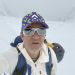 Man in a white jacket takes a selfie in a snowy, mountainous landscape wearing a purple-green cap and white sunglasses with a backpack behind him.
