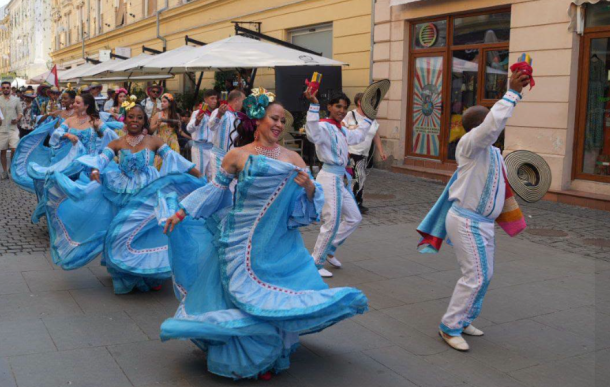 Group of dancers in flowing blue gowns performing a street parade, smiling and twirling feathers in the air against a city backdrop.