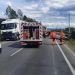 Emergency responders in orange vests at a highway accident scene with a tipped vehicle on the roadside and police/ambulance nearby