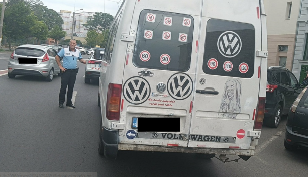 Rear view of a white Volkswagen van on a city street, covered with speed-limit stickers and decals, with a police officer standing nearby and other parked cars beside it.
