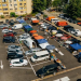 Overhead view of a bustling outdoor market in a parking lot, with colorful tents, vendors, and shoppers among parked cars and apartment buildings nearby.