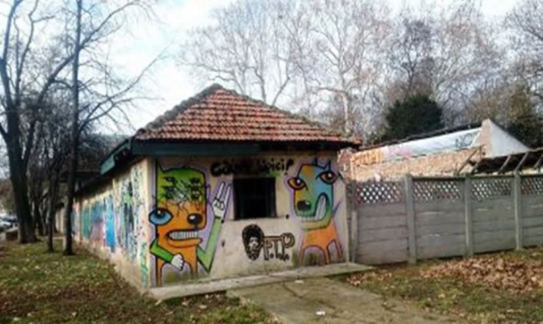 Small, graffiti-covered one-story building with a tiled, sloped roof and colorful cartoon faces on its walls, beside a path and fence.