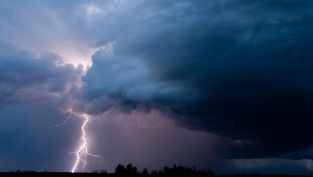 Lightning bolt streaks down through dark storm clouds over a flat horizon at dusk.
