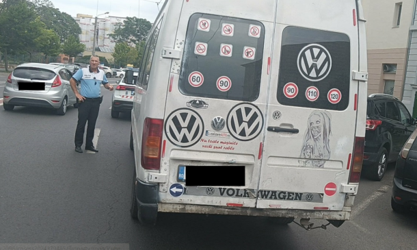 Rear view of a white Volkswagen van on a city street, covered with speed-limit stickers and decals, with a police officer standing nearby and other parked cars beside it.